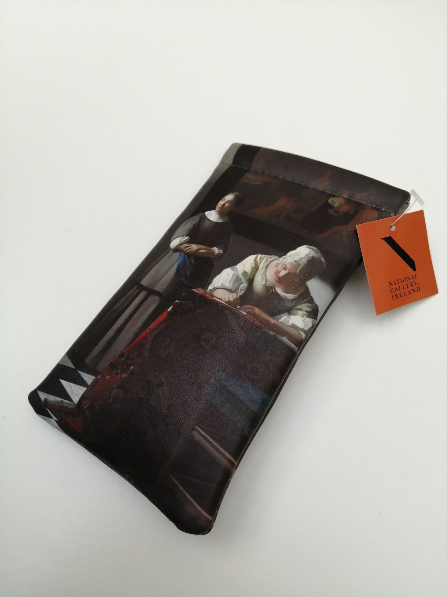 A rectangular pouch with an orange label. A woman stands looking off to the left. Beside her sitting at a table a woman is writing a letter.