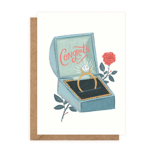 A white card with a drawing of an open blue ring box. Inside the lid ‘Congrats’ is written in red cursive. In the base is a gold diamond ring. A red rose is behind the box.