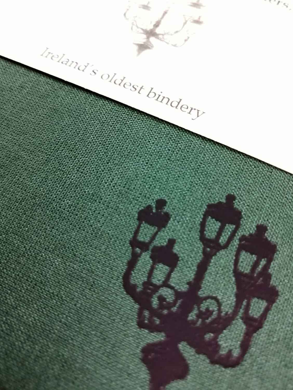 A close up of a lamp illustration on a dark green cloth notebook cover. A matching cream belly band is visible.